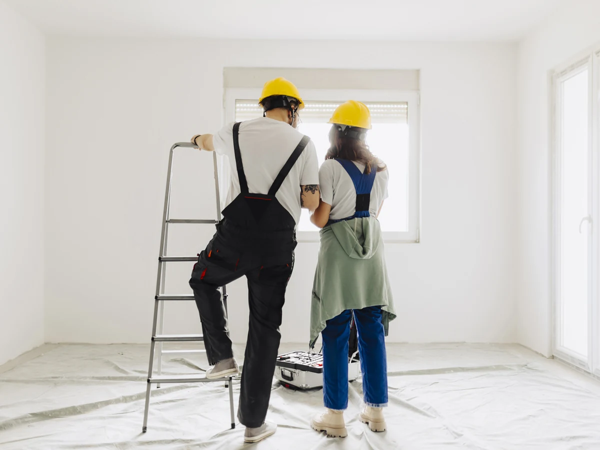 painting-decorating-two-workers-hard-york-painters Two decorators in yellow hard hats standing in an empty white room with dust sheets on the floor and a stepladder